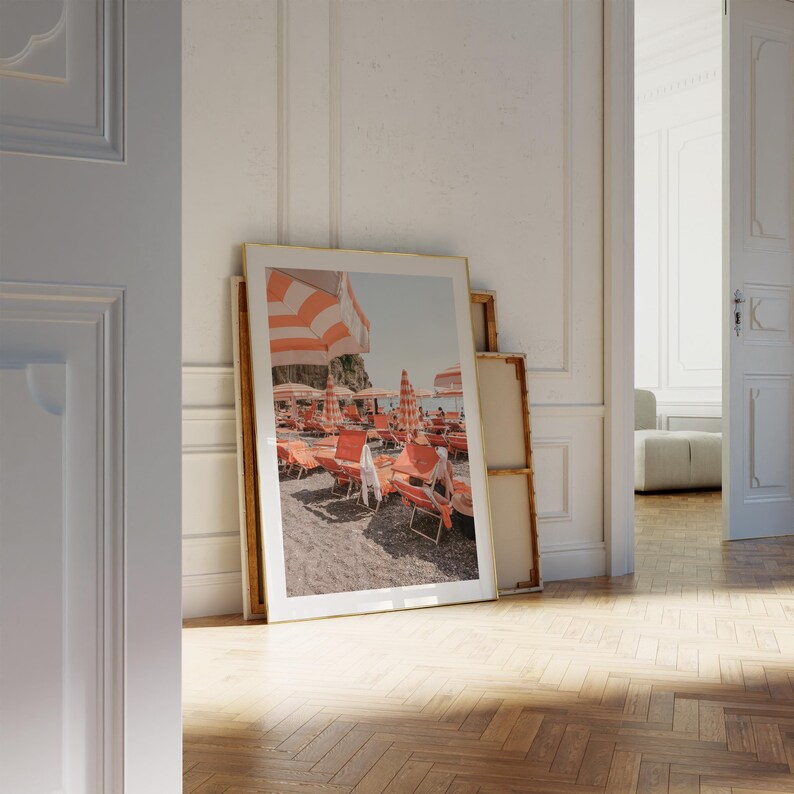 May include: A framed print of a beach scene with orange and white striped umbrellas and lounge chairs. The print is leaning against a wall in a room with a wooden floor. The image evokes a sunny, relaxing atmosphere.