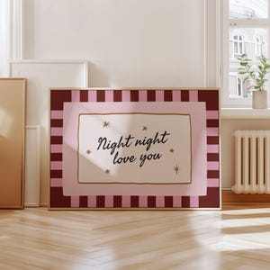 May include: A framed print with a pink and burgundy striped border, surrounding a light pink rectangle. The center features a cream-colored rectangle with the words "Night night love you" in a script font, with star accents.