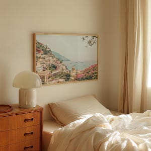 May include: A framed landscape print depicts a coastal Italian town with colorful buildings, mountains, and the sea. A white mushroom-shaped lamp sits on a wooden dresser. A bed with a beige pillow and white bedding is in the foreground.