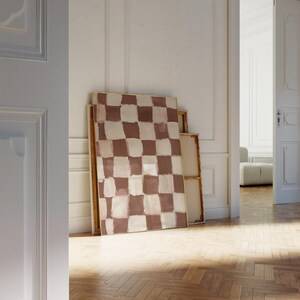 May include: A large abstract painting with a checkered pattern in shades of brown and white. The painting is framed in a thin gold frame and is leaning against a white wall.