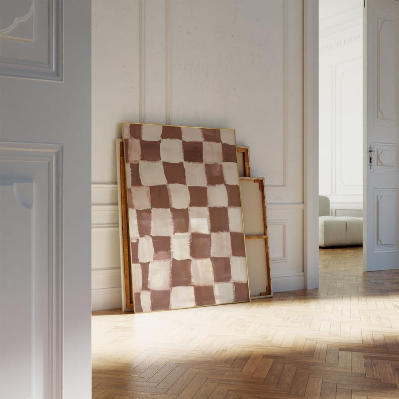 May include: A large abstract painting with a checkered pattern in shades of brown and white. The painting is framed in a thin gold frame and is leaning against a white wall.