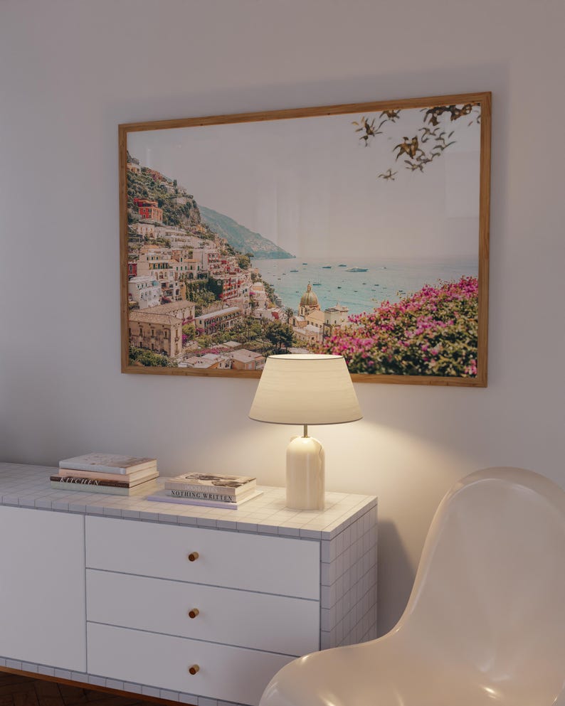 May include: A white dresser with three drawers and gold knobs. A lamp with a white shade sits on top of the dresser. A framed photograph of a coastal town with a blue ocean and pink flowers in the foreground is hanging on the wall behind the dresser.