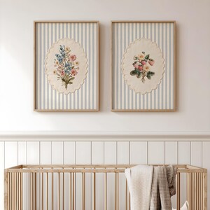 May include: Two framed floral prints. Each features a bouquet of flowers on an oval, scalloped-edge background, set against a striped backdrop. The frames are light wood. The bottom of the image shows a wooden crib with a blanket.