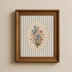 K&ouml;nnte beinhalten: Gerahmte Blumenstickerei mit einem Bouquet aus rosa, blauen und gelben Blumen. Die Stickerei befindet sich vor einem gestreiften Hintergrund aus hellblau und wei&szlig;, in einem Holzrahmen.