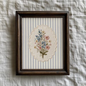 K&ouml;nnte beinhalten: Gerahmte Blumenstickerei mit einem Bouquet aus rosa, blauen und gelben Blumen. Die Stickerei befindet sich vor einem gestreiften Hintergrund aus hellblau und wei&szlig;, in einem dunkelbraunen Holzrahmen.