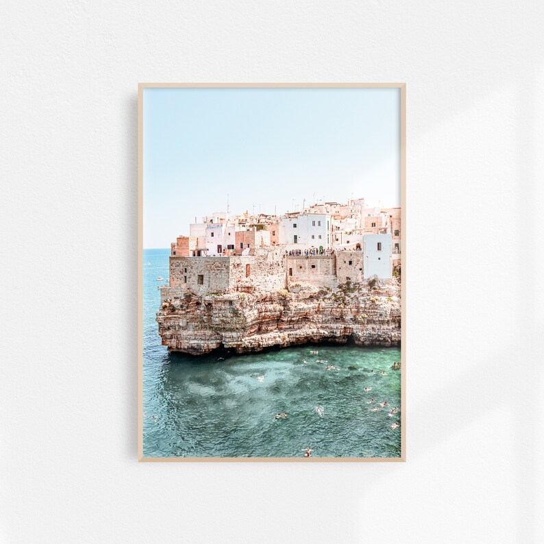 May include: A photograph of a white and beige building on a cliff overlooking a turquoise body of water. People are swimming in the water.