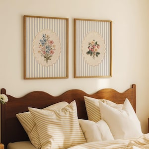 May include: Two framed floral art prints. Each features an oval-shaped embroidered floral design on a striped background of light blue and white. The frames are a warm, light brown color. The prints are displayed above a bed with striped pillows and bedding.