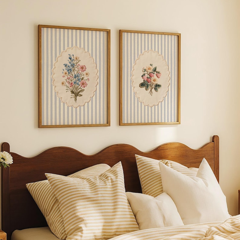 May include: Two framed floral art prints. Each features an oval-shaped embroidered floral design on a striped background of light blue and white. The frames are a warm, light brown color. The prints are displayed above a bed with striped pillows and bedding.