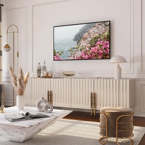 May include: A white cabinet with gold accents and a white marble top. The cabinet has a textured, ribbed design. A lamp with a white shade sits on top of the cabinet. A large framed photograph of a coastal scene with pink flowers hangs on the wall above the cabinet. A white vase with dried pampas grass sits on the floor next to the cabinet. A white marble coffee table sits in front of the cabinet. A brown velvet stool with a gold metal frame sits in front of the coffee table.