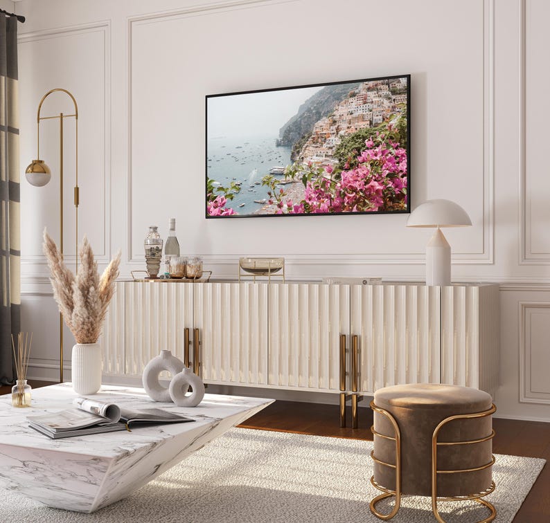 May include: A white cabinet with gold accents and a white marble top. The cabinet has a textured, ribbed design. A lamp with a white shade sits on top of the cabinet. A large framed photograph of a coastal scene with pink flowers hangs on the wall above the cabinet. A white vase with dried pampas grass sits on the floor next to the cabinet. A white marble coffee table sits in front of the cabinet. A brown velvet stool with a gold metal frame sits in front of the coffee table.