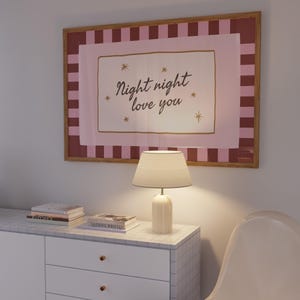 May include: A framed art print with the words "Night night love you" in a cursive font, surrounded by stars. The print is above a white cabinet with a lamp and books. The frame has a pink and brown striped border.
