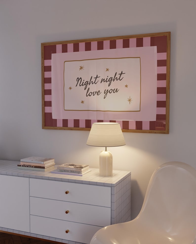 May include: A framed art print with the words "Night night love you" in a cursive font, surrounded by stars. The print is above a white cabinet with a lamp and books. The frame has a pink and brown striped border.