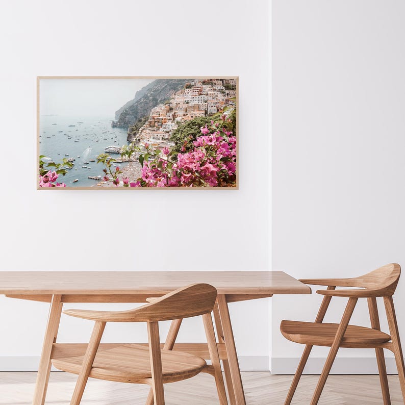 May include: A framed photograph of a coastal town with colorful buildings on a cliffside overlooking a blue ocean. Pink flowers are in the foreground.