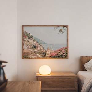 May include: A framed landscape print of a coastal Italian town hangs above a wooden nightstand. The print features colorful buildings on a hillside, with a body of water in the background. A glowing, spherical lamp sits on the nightstand.