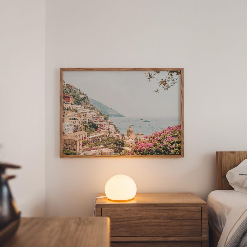 May include: A framed landscape print of a coastal Italian town hangs above a wooden nightstand. The print features colorful buildings on a hillside, with a body of water in the background. A glowing, spherical lamp sits on the nightstand.