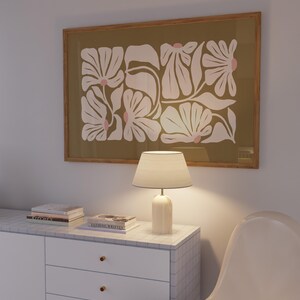 May include: A framed floral art print with white flowers and pink accents on an olive green background. A white lamp with a beige lampshade sits on a white cabinet with a marble top. The cabinet has three drawers with gold knobs.