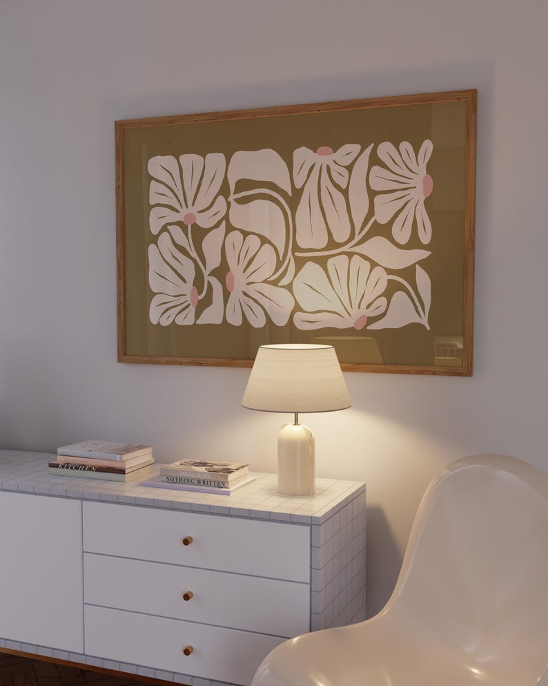 May include: A framed floral art print with white flowers and pink accents on an olive green background. A white lamp with a beige lampshade sits on a white cabinet with a marble top. The cabinet has three drawers with gold knobs.