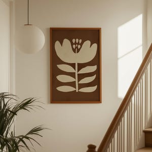 Pu&ograve; includere: Una stampa artistica incorniciata con un fiore bianco stilizzato su sfondo marrone. L'opera d'arte &egrave; in una cornice di legno e appesa a una parete bianca. Una lampada a globo bianca pende dal soffitto.