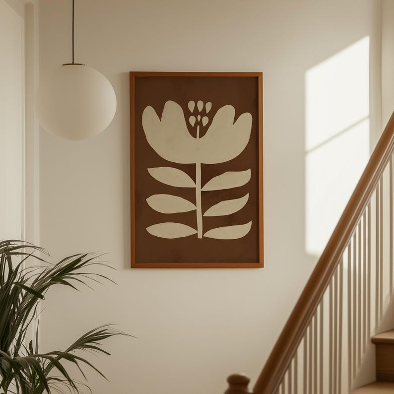 Pu&ograve; includere: Una stampa artistica incorniciata con un fiore bianco stilizzato su sfondo marrone. L'opera d'arte &egrave; in una cornice di legno e appesa a una parete bianca. Una lampada a globo bianca pende dal soffitto.