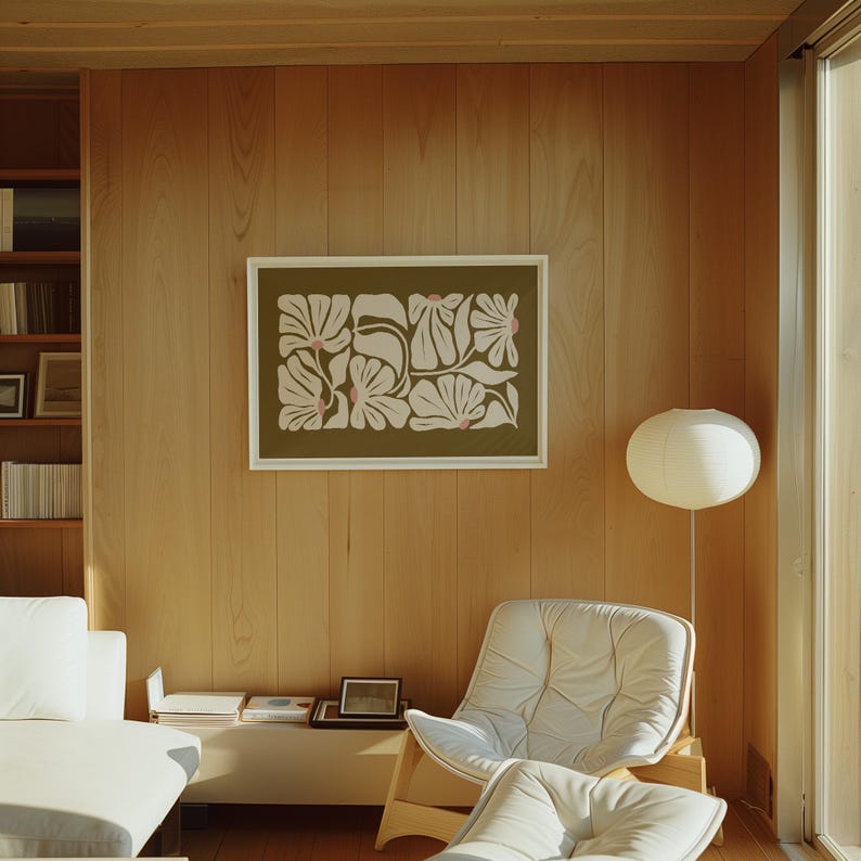May include: A framed floral art print with a white border hangs on a light wood-paneled wall. The artwork features a repeating pattern of stylized white flowers with pink accents on an olive green background. A white chair and a floor lamp are also visible.