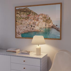 May include: A white dresser with three drawers and gold knobs. A lamp with a white shade sits on top of the dresser. A framed photograph of a coastal town with colorful buildings on a cliffside overlooking a blue ocean is hanging on the wall behind the dresser.
