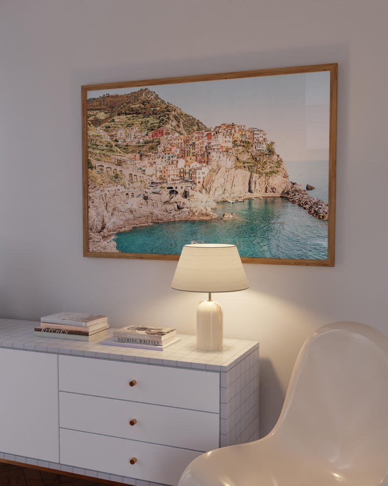 May include: A white dresser with three drawers and gold knobs. A lamp with a white shade sits on top of the dresser. A framed photograph of a coastal town with colorful buildings on a cliffside overlooking a blue ocean is hanging on the wall behind the dresser.
