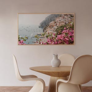 May include: A framed photograph of a coastal town with colorful buildings, lush greenery, and boats in the water. The photo is framed in a light wood frame and is hanging on a light beige wall.