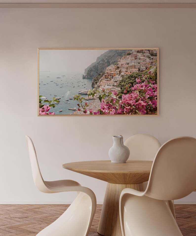 May include: A framed photograph of a coastal town with colorful buildings, lush greenery, and boats in the water. The photo is framed in a light wood frame and is hanging on a light beige wall.