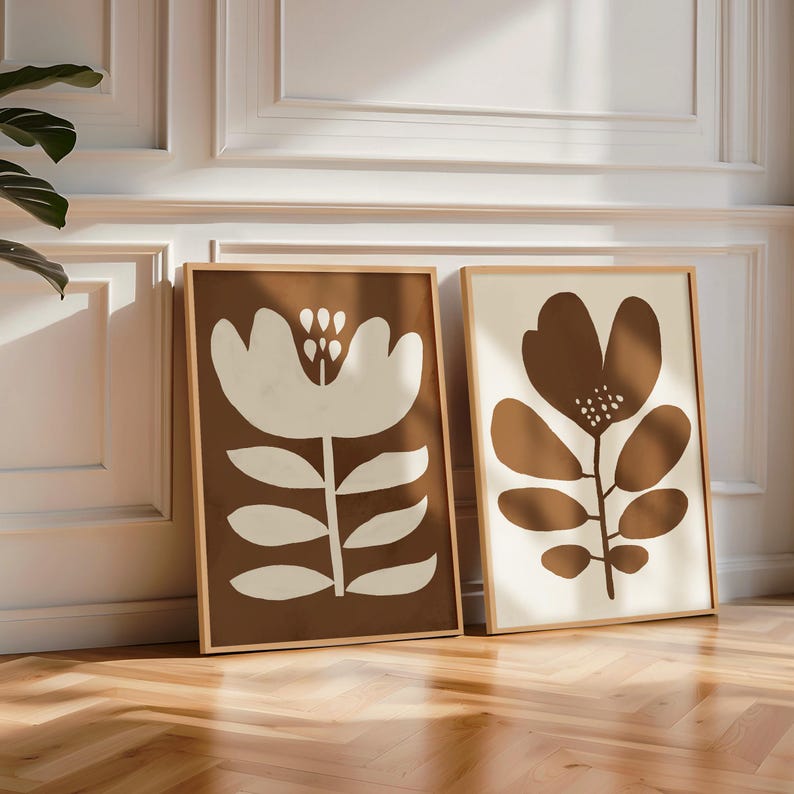 Pu&ograve; includere: Due stampe artistiche floreali incorniciate. Una stampa presenta un grande fiore bianco su sfondo marrone. L'altra ha un fiore marrone su sfondo crema. Entrambe sono in cornici di legno e appoggiate a un muro bianco.