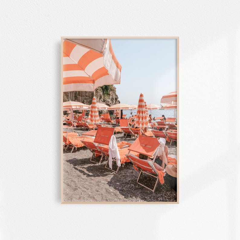 May include: A framed print of a beach scene with orange and white striped umbrellas and matching lounge chairs. The beach is filled with people enjoying the sun and the ocean. The sky is a clear blue.