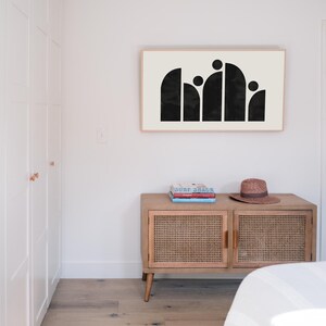 May include: A black and white abstract art print in a wooden frame hangs on a white wall above a wooden cabinet with a woven rattan front. The cabinet has two doors and is sitting on four legs. There are three books stacked on top of the cabinet.