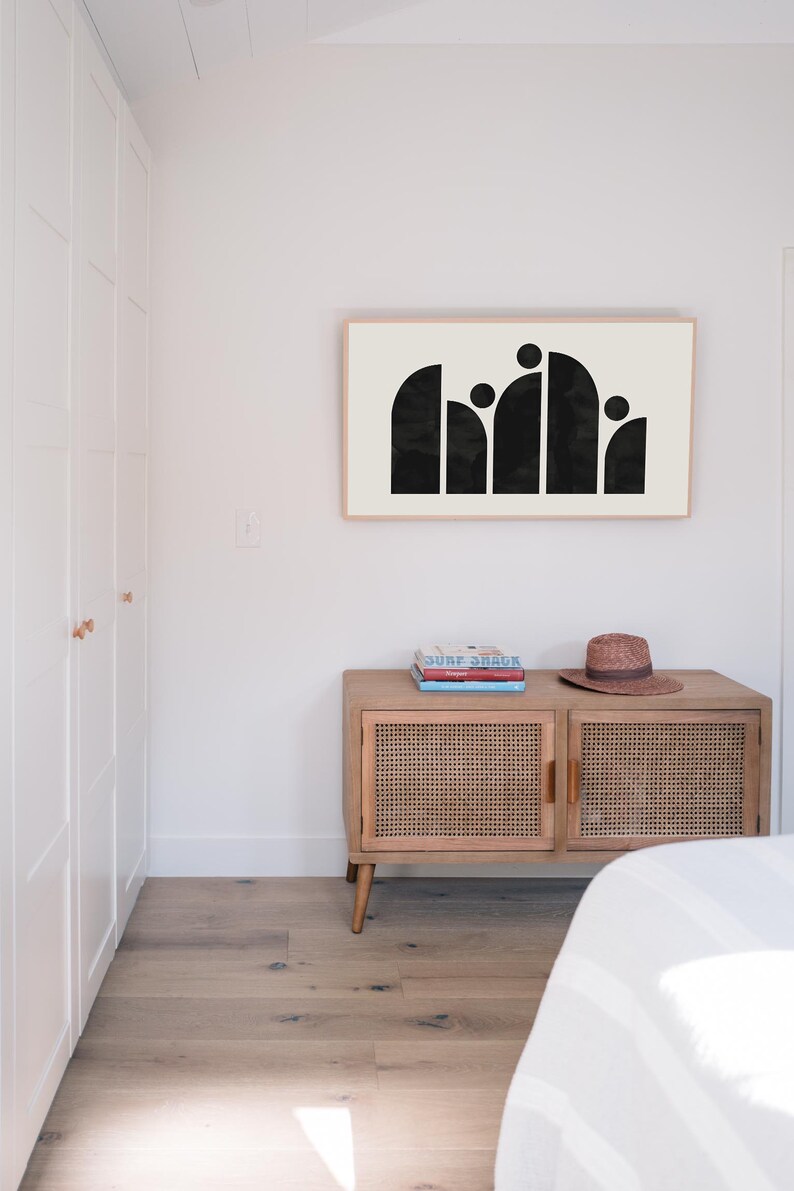 May include: A black and white abstract art print in a wooden frame hangs on a white wall above a wooden cabinet with a woven rattan front. The cabinet has two doors and is sitting on four legs. There are three books stacked on top of the cabinet.