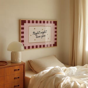 May include: A framed wall art print with the words "Night night love you" in a bedroom setting. The artwork has a pink and burgundy border with a cream-colored center. A white mushroom lamp sits on a wooden nightstand.