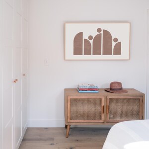 May include: A light brown abstract art print with a white background hangs on a white wall above a wooden cabinet with a woven rattan front. A straw hat sits on top of the cabinet, which has three books stacked on top of it.