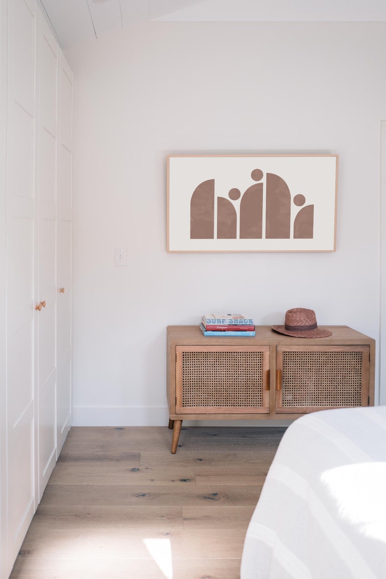 May include: A light brown abstract art print with a white background hangs on a white wall above a wooden cabinet with a woven rattan front. A straw hat sits on top of the cabinet, which has three books stacked on top of it.