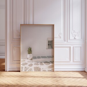 May include: A framed print featuring a serene outdoor scene. The image shows a white wall with a small window, a cactus in a white pot, and a stone pathway. The frame is a light wood color, and the room has a hardwood floor.
