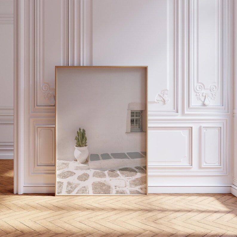 May include: A framed print featuring a serene outdoor scene. The image shows a white wall with a small window, a cactus in a white pot, and a stone pathway. The frame is a light wood color, and the room has a hardwood floor.