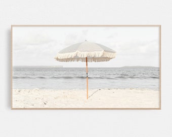 May include: A white and blue striped beach umbrella with a fringe border stands on a sandy beach with the ocean in the background.