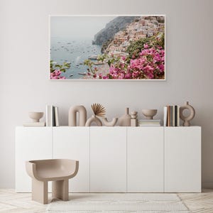 May include: A framed photograph of a coastal town with colorful buildings built into a cliffside. The town is surrounded by a blue ocean with boats and a lush green hillside. Pink flowers are in the foreground.