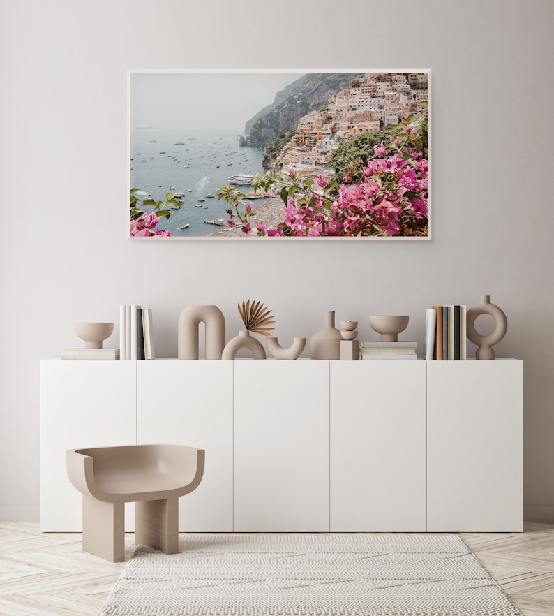 May include: A framed photograph of a coastal town with colorful buildings built into a cliffside. The town is surrounded by a blue ocean with boats and a lush green hillside. Pink flowers are in the foreground.