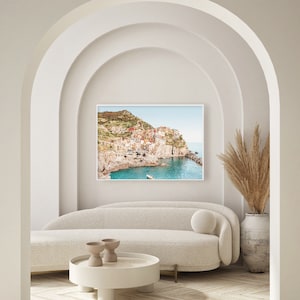 May include: A white couch with a round pillow sits in front of a framed photograph of a colorful Italian village on a cliff overlooking the ocean. A vase with dried pampas grass sits on the right side of the image.