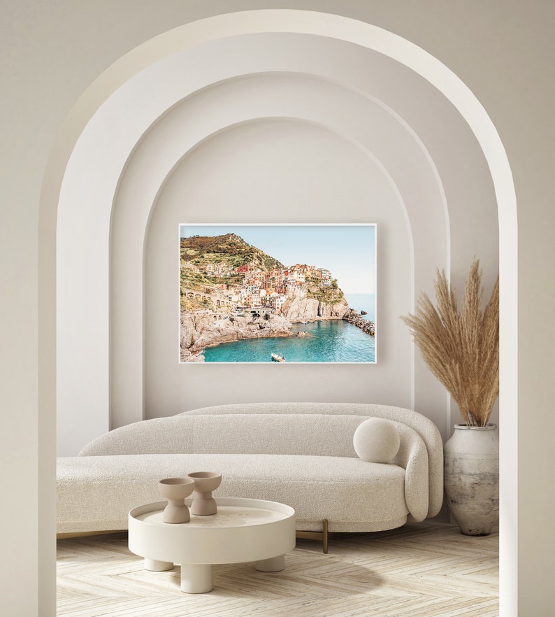 May include: A white couch with a round pillow sits in front of a framed photograph of a colorful Italian village on a cliff overlooking the ocean. A vase with dried pampas grass sits on the right side of the image.