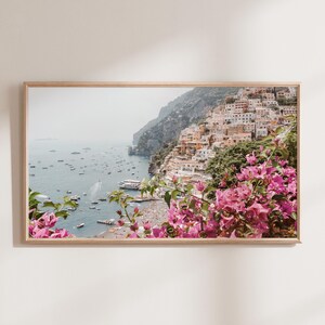 May include: A scenic photograph of a coastal town in Italy, with colorful buildings built into the hillside. The town is nestled between a lush green hillside and the sparkling blue sea. Pink flowers bloom in the foreground, framing the view.