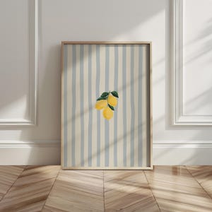 May include: A framed art print featuring a watercolor illustration of three lemons with green leaves. The background has vertical stripes in light blue and cream colors. The frame is made of light-colored wood.