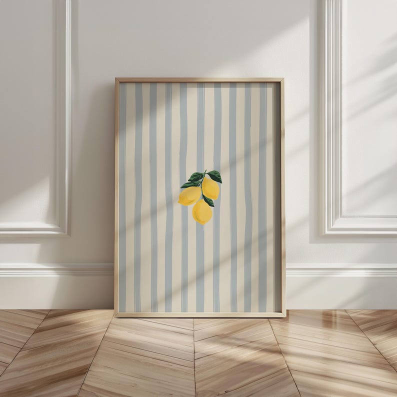 May include: A framed art print featuring a watercolor illustration of three lemons with green leaves. The background has vertical stripes in light blue and cream colors. The frame is made of light-colored wood.