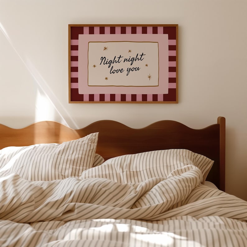 May include: A framed print with the text "Night night love you" in a bedroom setting. The print has a pink and burgundy color scheme with a gold frame. The bed has striped bedding and a wooden headboard.