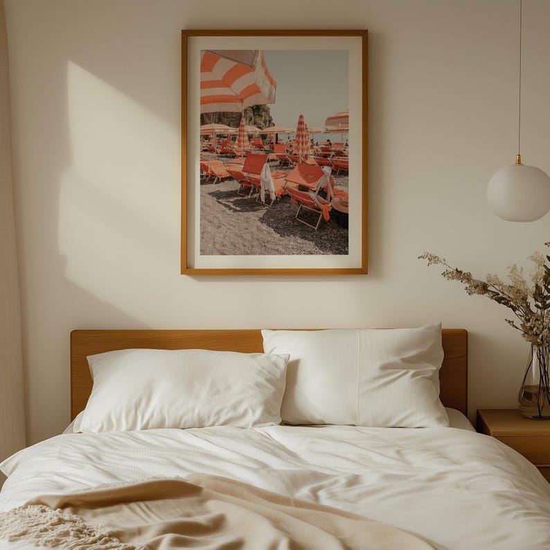 May include: A framed print depicts a beach scene with orange and white striped umbrellas and lounge chairs. The print is in a wooden frame and hangs on a cream-colored wall above a bed with white bedding.