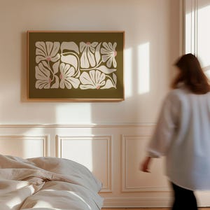 May include: A framed floral art print featuring a botanical design with white and pink flowers on an olive green background. The artwork is displayed on a white wall in a bedroom setting, with a bed and a person in the background.