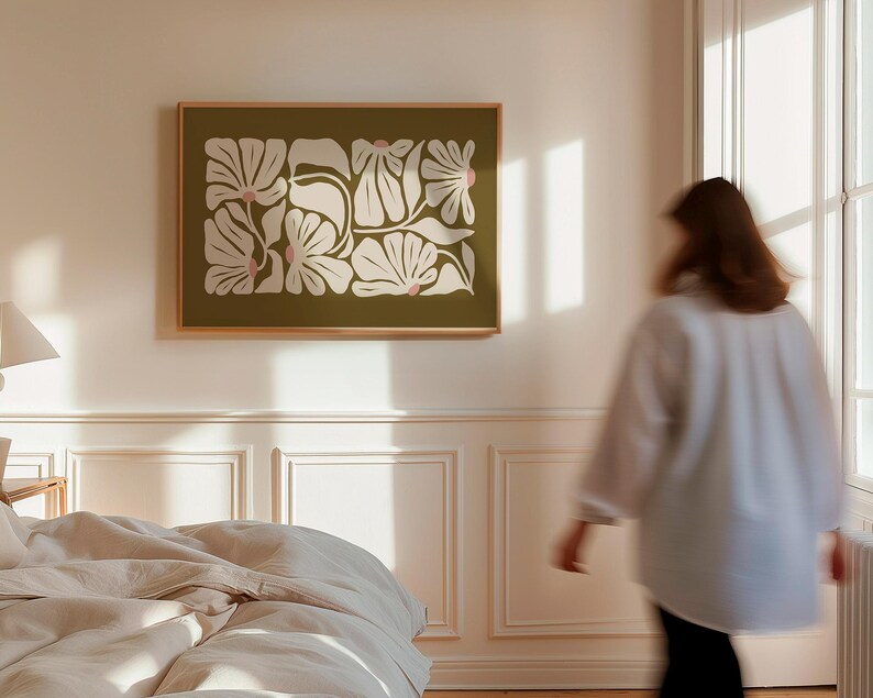 May include: A framed floral art print featuring a botanical design with white and pink flowers on an olive green background. The artwork is displayed on a white wall in a bedroom setting, with a bed and a person in the background.