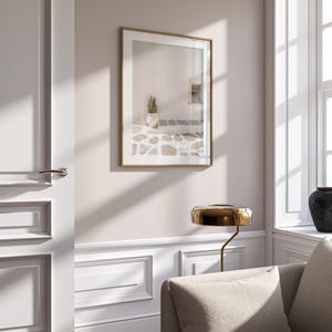 May include: A framed print with a gold-colored frame hangs on a light pink wall. The print depicts a white building with a cactus in a white pot. A gold-colored lamp and a beige sofa are in the foreground.